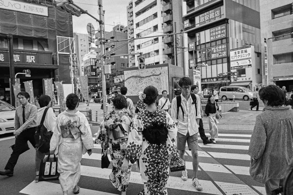 Black and white photo of group of people crossing crosswalk in urban Japan, some wearing kimonos. Buildings and signs in background.