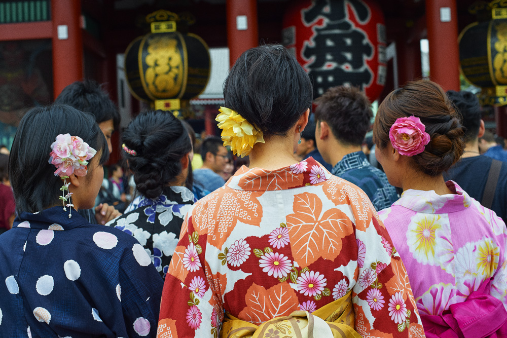 Group of people wearing colourful traditional yukata robes with floral hair ornaments seen from behind at a shrine.