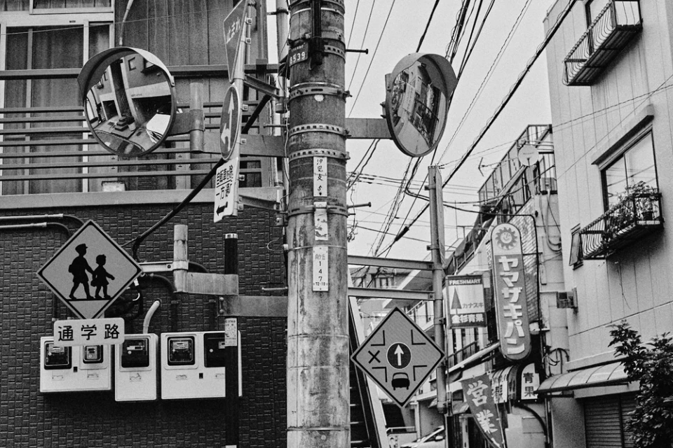 Monochrome image of a street scene with utility pole, signs, electrical boxes, and buildings in black and white tones.