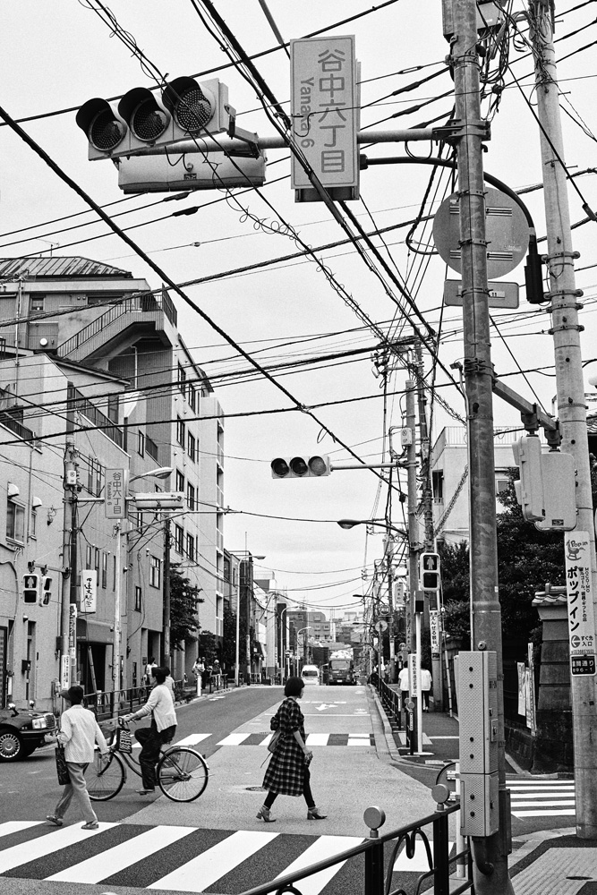 Black and white street scene in Japan featuring pedestrians crossing a crosswalk, utility poles, and buildings, capturing urban life.