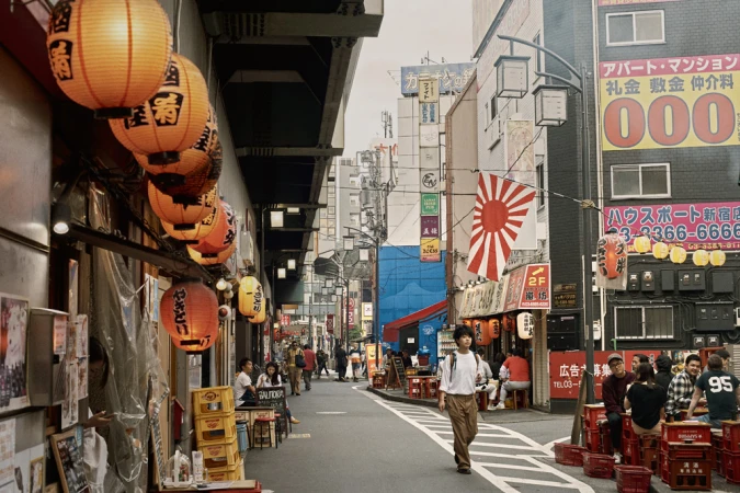 A person walking down a busy narrow street in Japan lined with glowing orange paper lanterns and outdoor dining tables.