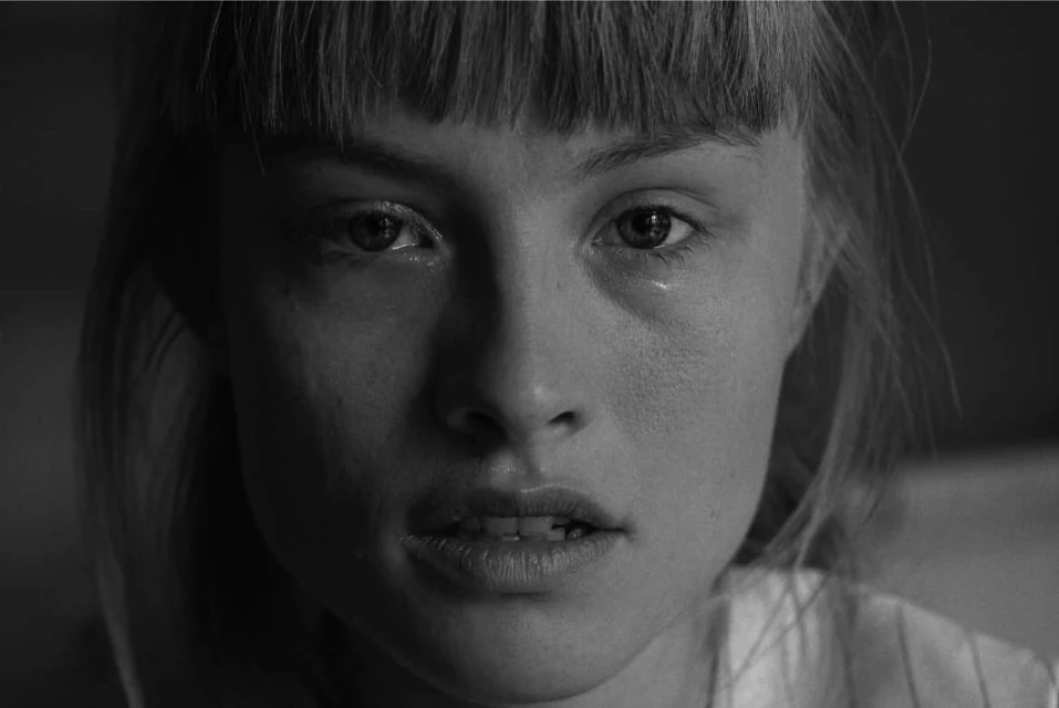 Close-up black and white portrait of a young adult woman with bangs, looking directly at the camera with a tear on her face.
