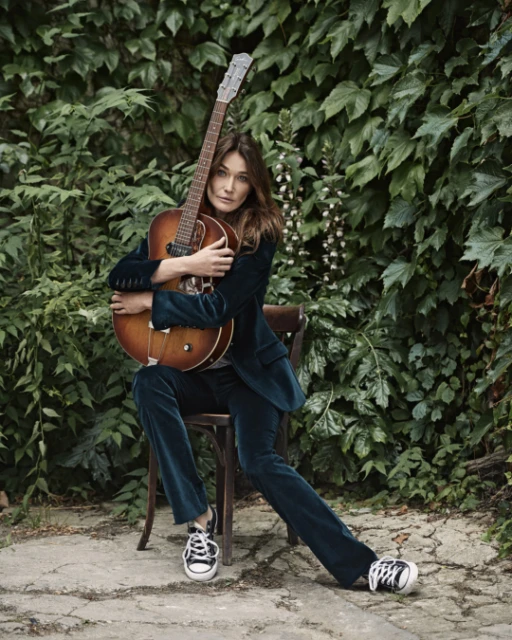 Adult female musician poses with a guitar, seated outdoors, in a green velvet suit and sneakers, surrounded by lush foliage.