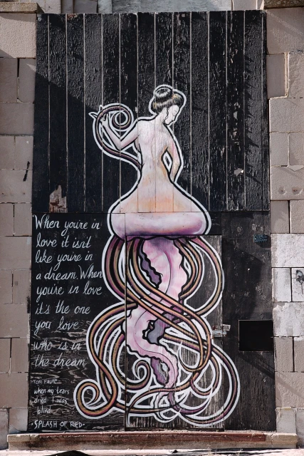 Illustration on a weathered door showing a nude figure with flowing lines and script, creating an artistic mural on an urban backdrop.