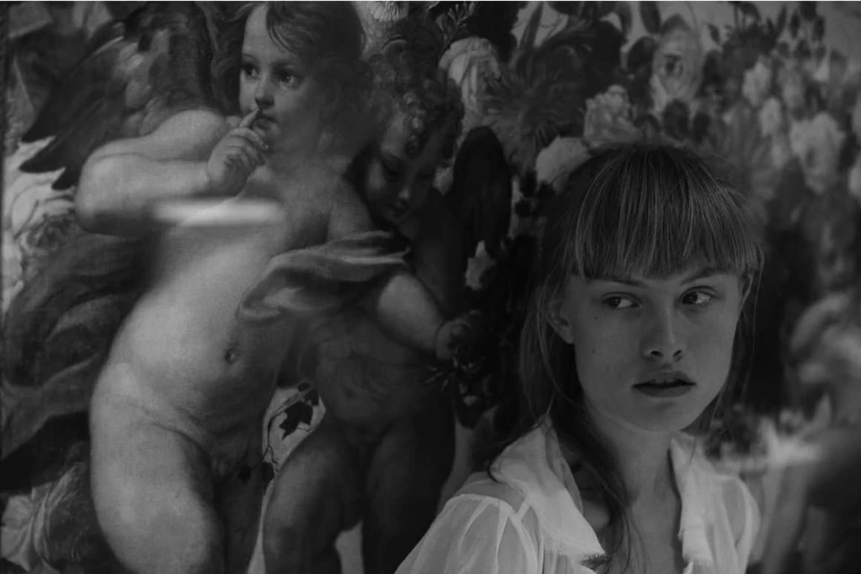Black and white portrait of a young woman with bangs, standing in front of a floral patterned wall and a faded cherub painting.