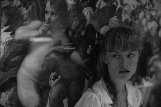 Black and white portrait of a young woman with bangs, standing in front of a floral patterned wall and a faded cherub painting.