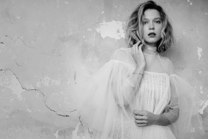 Black and white portrait of a woman wearing a sheer tulle high neck dress posing against a distressed wall.