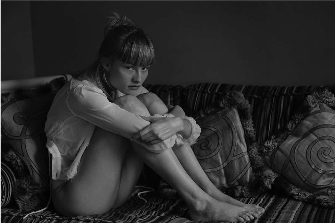 Young woman with bangs sits on sofa, hugging her legs, looking at the camera in black and white tones, captured in an indoor setting.