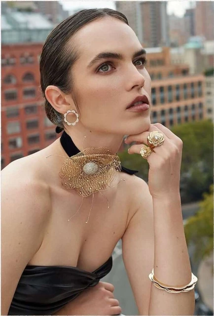 A woman wearing jewelry poses outdoors in a black top with a city building background, showcasing fashion and beauty.