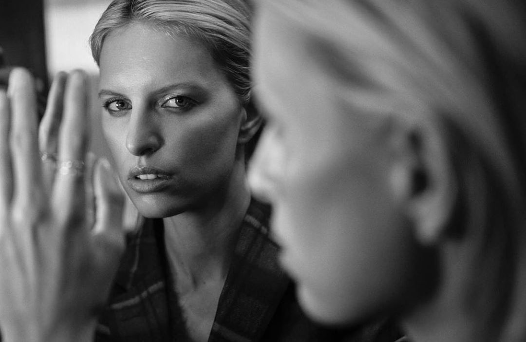 Black and white portrait of a blonde woman reflected in a mirror with her hand raised, captured by Eric Guillemain.