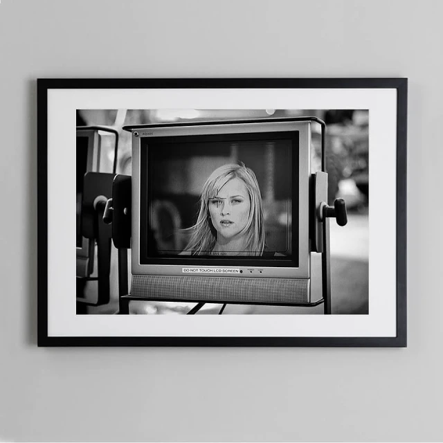 Black and white photograph of a television monitor displaying a woman with blonde hair and a serious expression, set against an outdoor background.