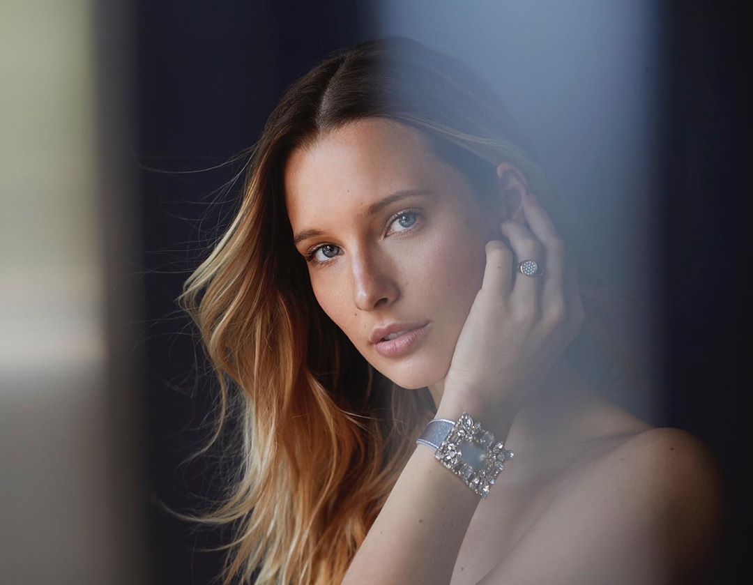 Close-up portrait of a blonde woman with a neutral expression, wearing a sparkling ring and a jeweled bracelet against a dark background.