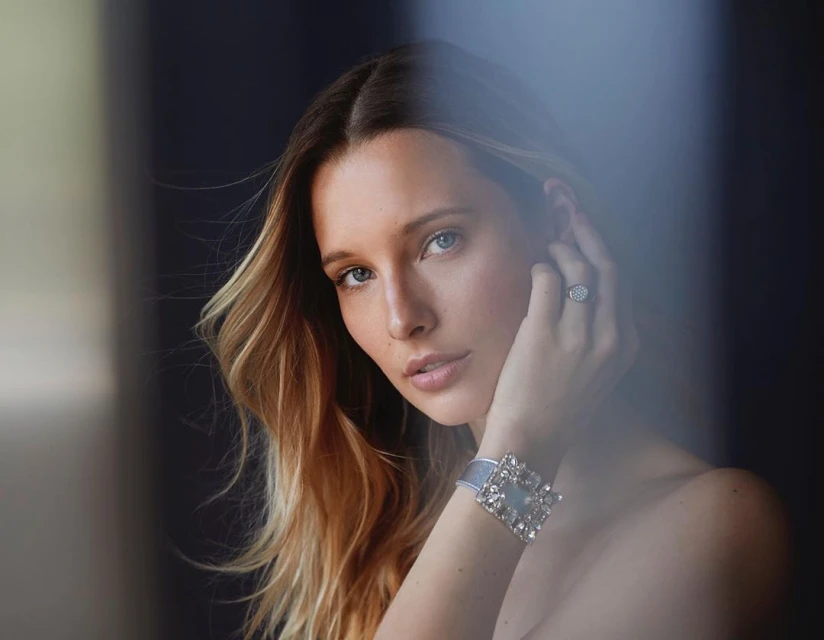 Close-up portrait of a blonde woman with a neutral expression, wearing a sparkling ring and a jeweled bracelet against a dark background.
