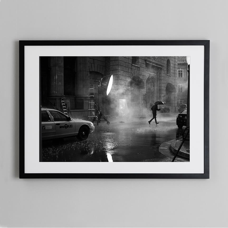 Black and white photograph capturing a person crossing a street under an umbrella in the rain with a taxi, lighting equipment, and buildings.