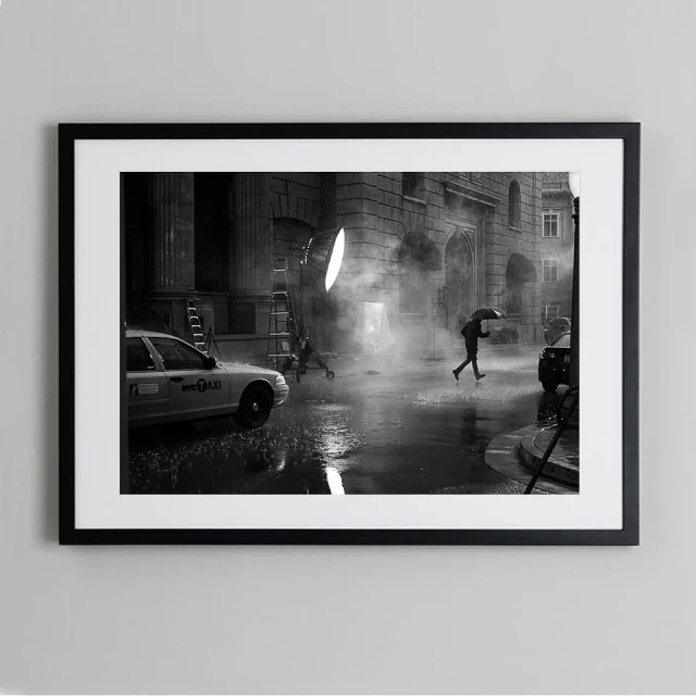 Black and white photograph capturing a person crossing a street under an umbrella in the rain with a taxi, lighting equipment, and buildings.