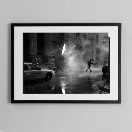 Black and white photograph capturing a person crossing a street under an umbrella in the rain with a taxi, lighting equipment, and buildings.