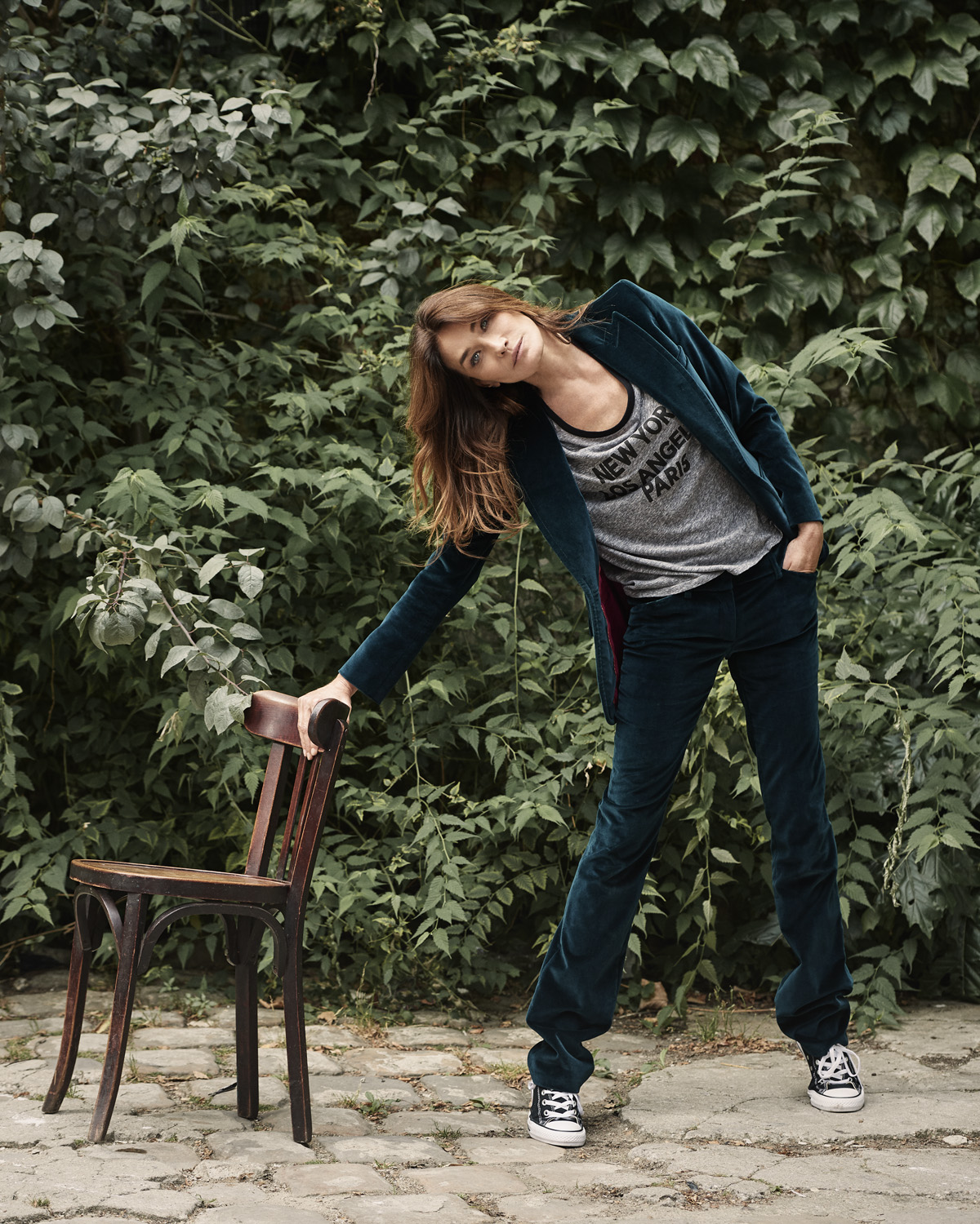 An adult woman in a green velvet suit poses leaning against a wooden chair outdoors, with lush green foliage filling the background.