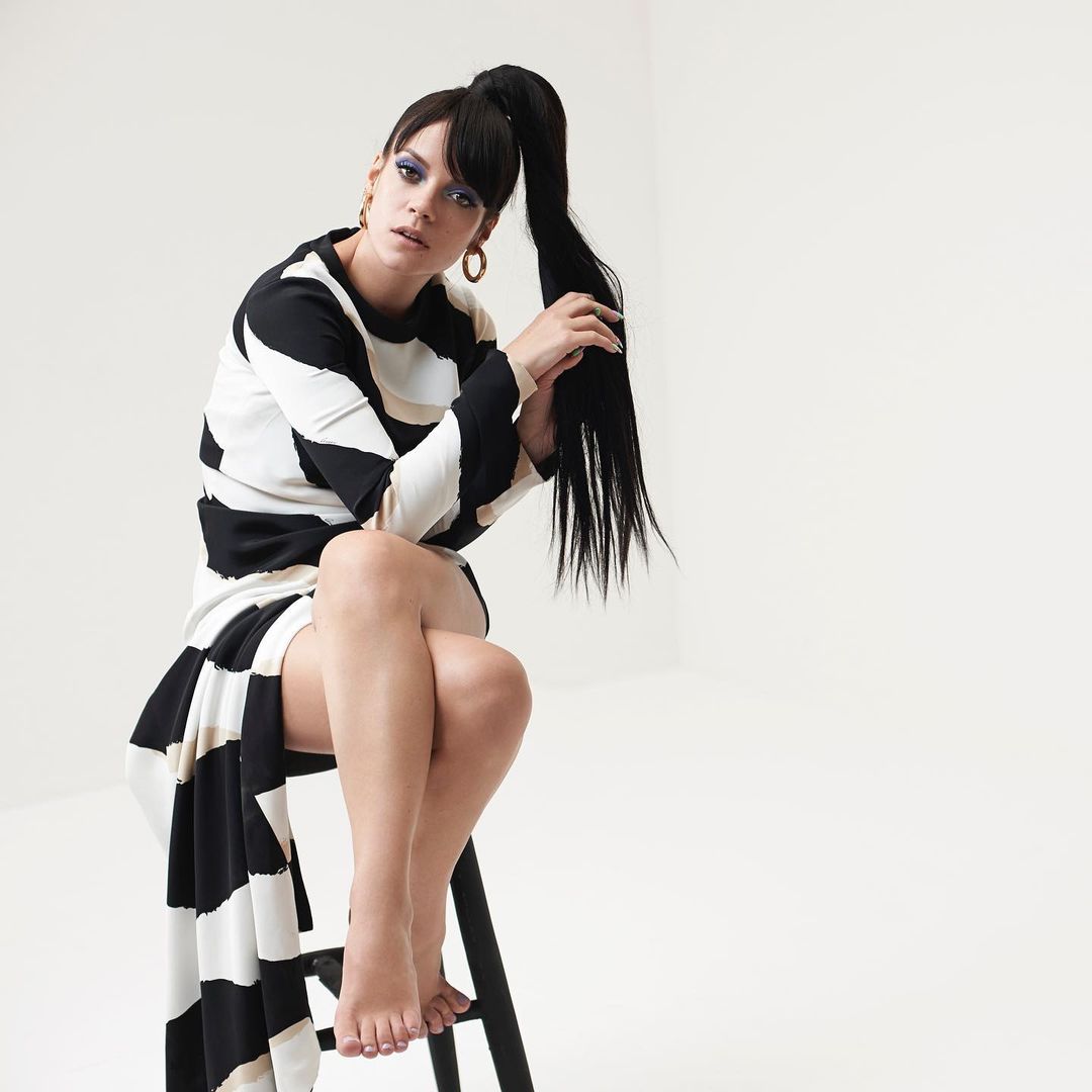Woman with long black high ponytail holding her hair while seated on a stool wearing a black and white patterned dress