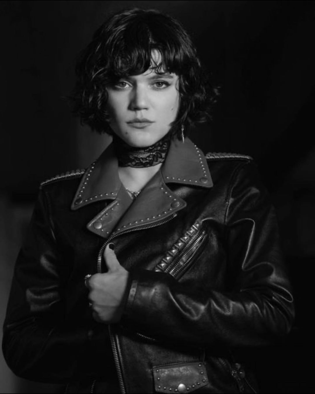 Monochrome portrait of a woman with dark wavy hair wearing a studded leather jacket and a lace choker indoors, facing forward.