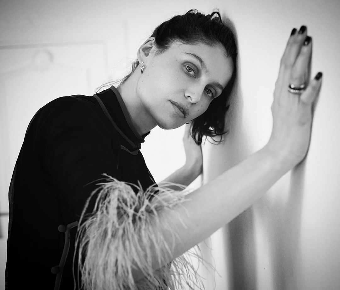 Monochrome portrait of a young woman leaning against a white wall, wearing a dark top with feather details, indoors with natural lighting.
