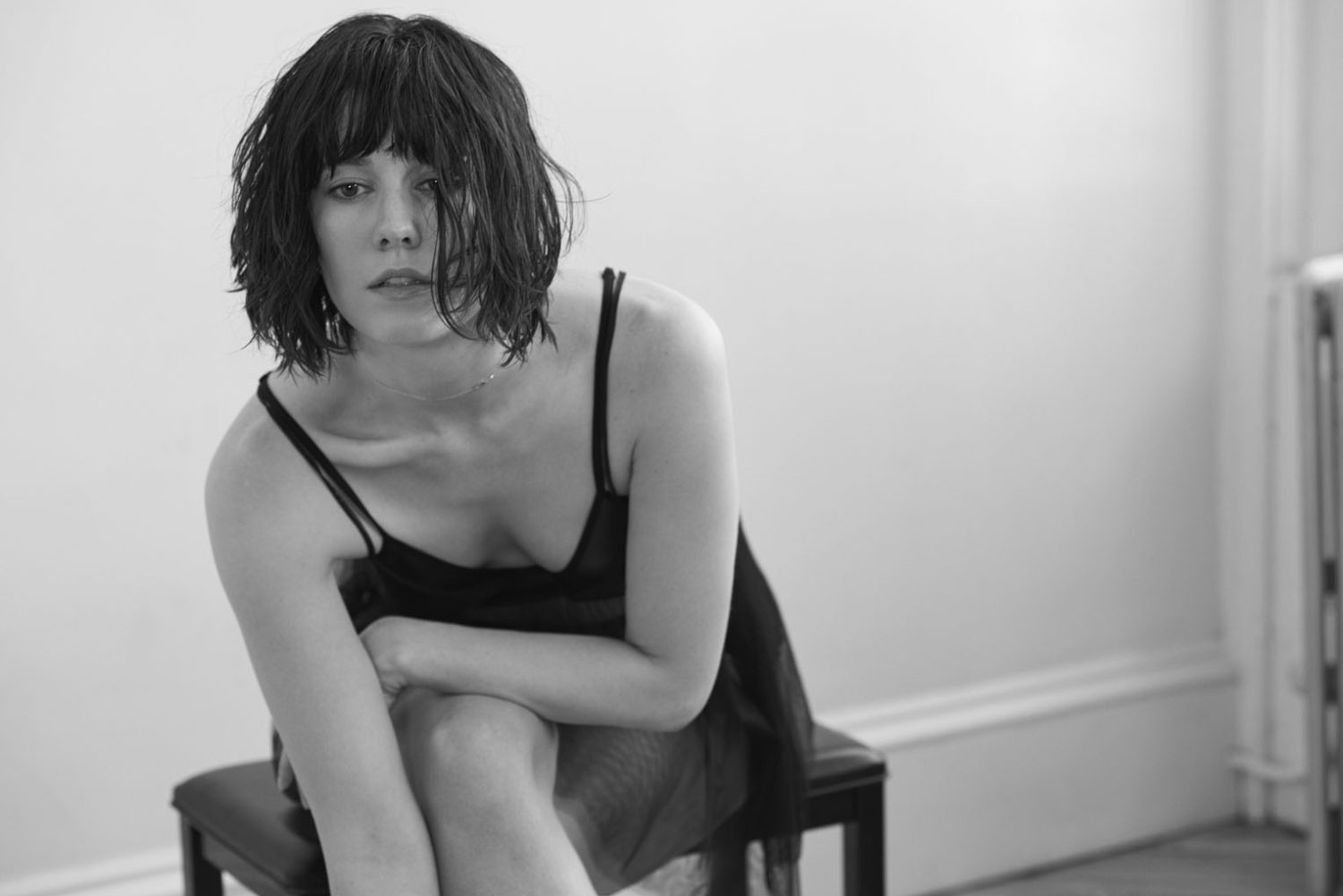 Black and white portrait of a female adult with short dark hair, sitting on a stool in front of a white wall in a natural setting.