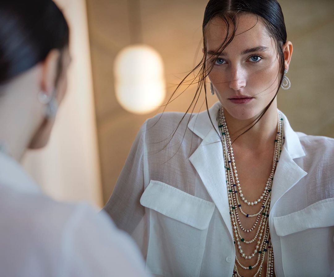 Woman in a white shirt and layered pearl necklaces gazes intensely, reflected in a mirror. Soft lighting enhances the serene scene.