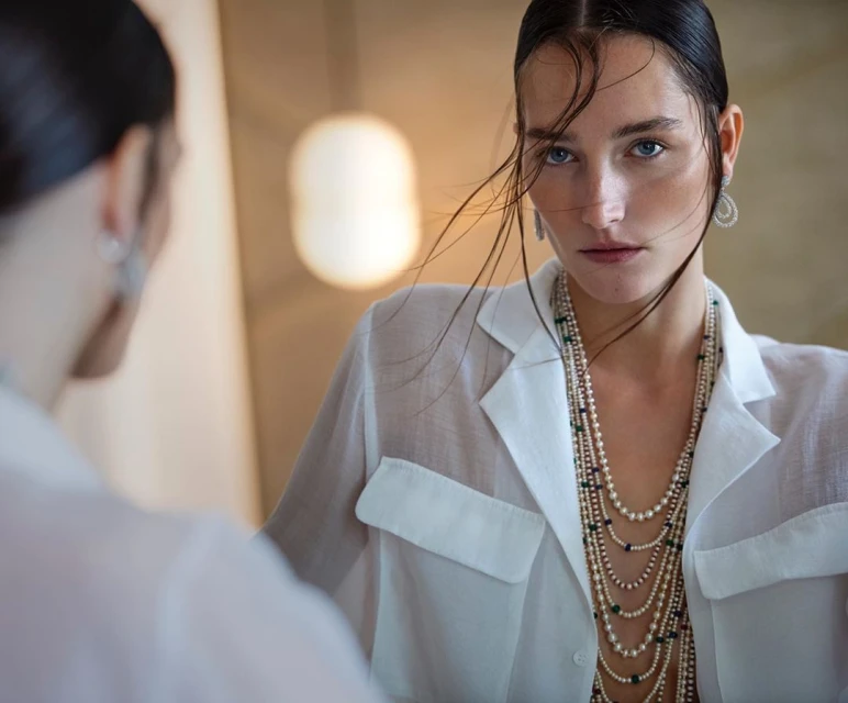 Woman in a white shirt and layered pearl necklaces gazes intensely, reflected in a mirror. Soft lighting enhances the serene scene.