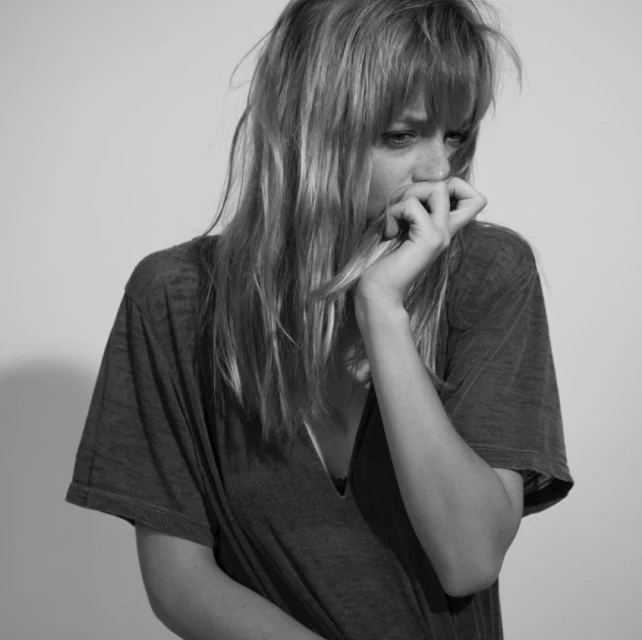 Monochrome portrait of a thoughtful young woman with bangs, wearing a dark V-neck shirt, hand near face, against a light grey backdrop.