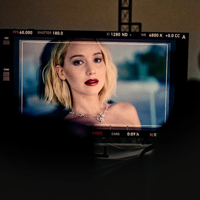 Jennifer Lawrence close-up on film set monitor, fair skin, blonde bob, dark red lips, bare shoulder, diamond necklace, cool light, indoor.