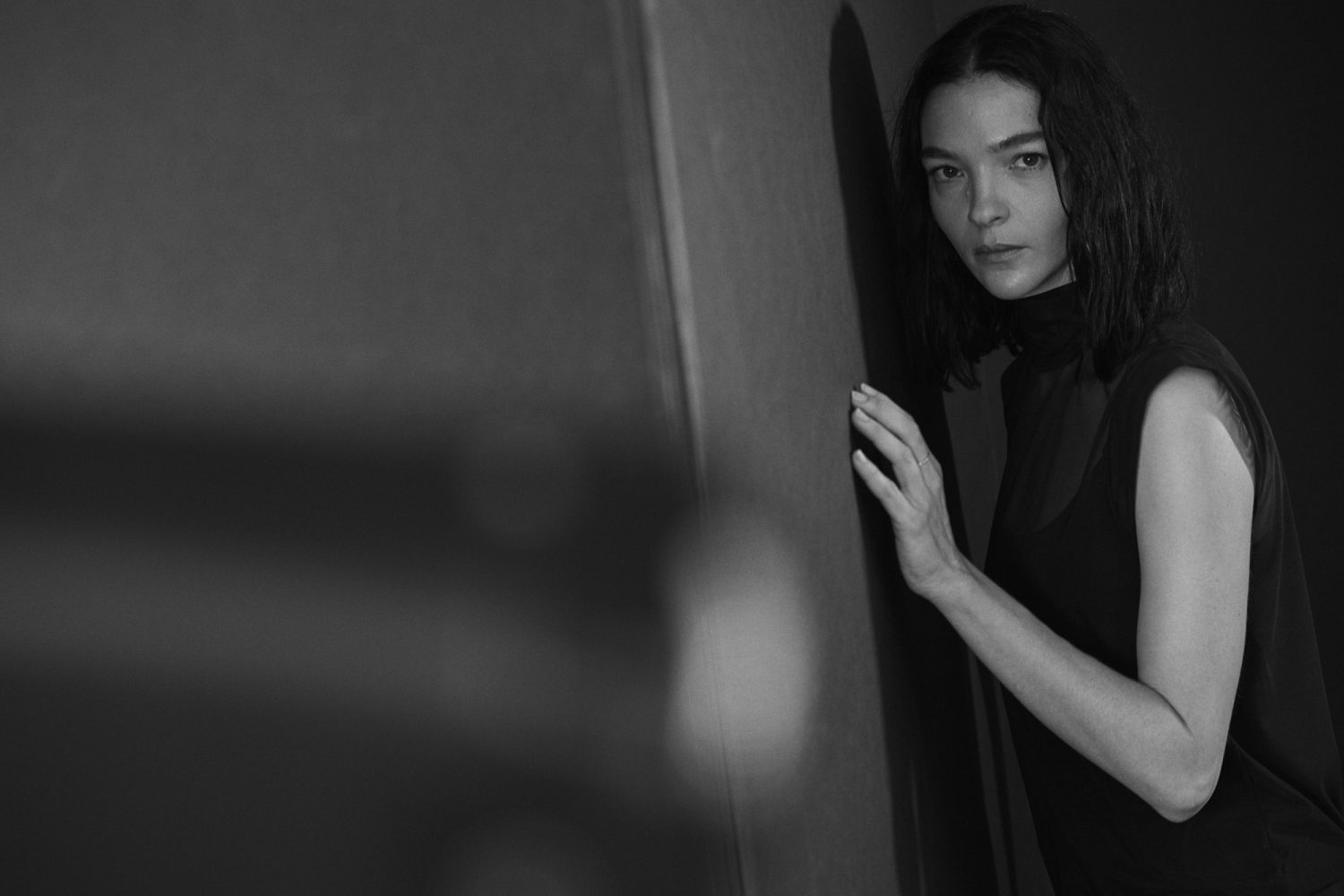 Monochrome portrait of a pensive young woman in a black top leaning against a wall, with soft lighting and a minimalist aesthetic.