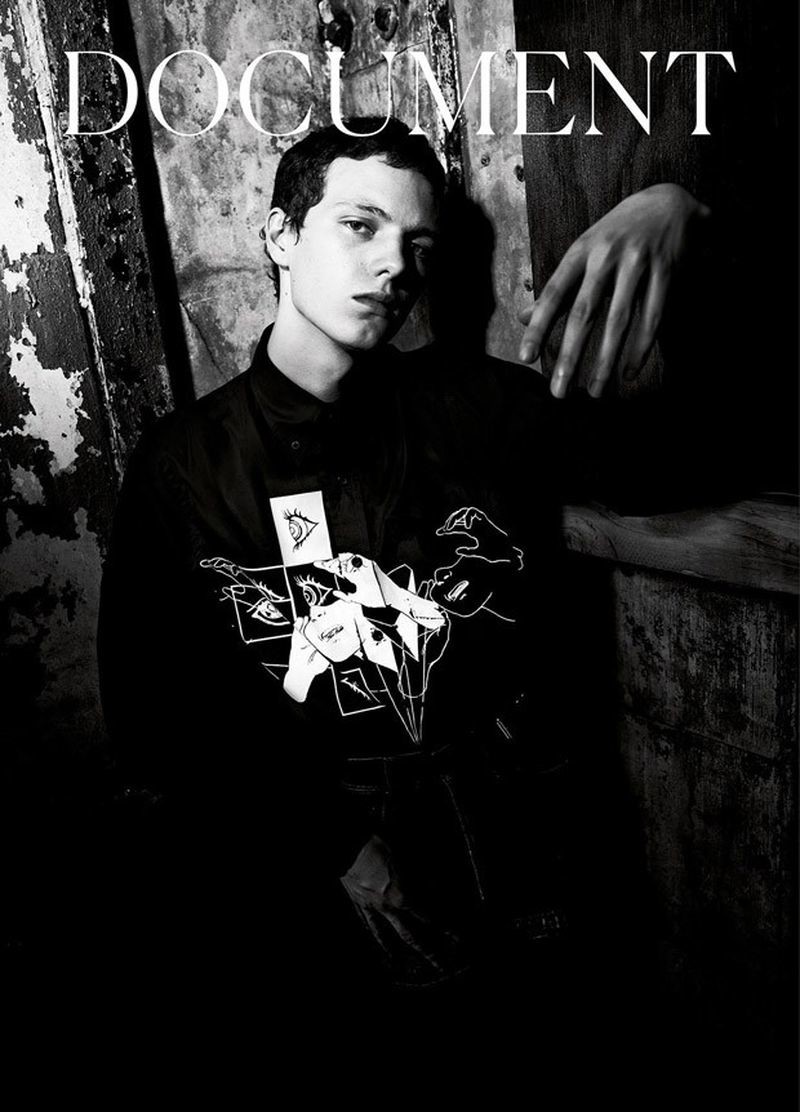 Black-and-white fashion portrait of a young adult male model wearing a graphic top, posing in front of a textured wall with hand visible.