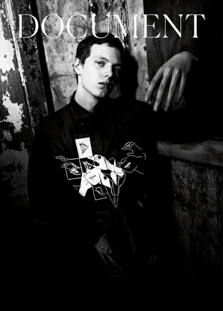 Black-and-white fashion portrait of a young adult male model wearing a graphic top, posing in front of a textured wall with hand visible.