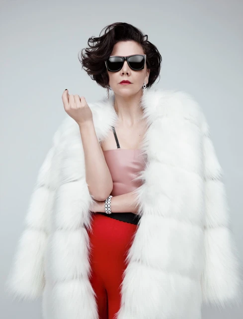 Stylish woman in a white faux fur coat and sunglasses poses against a neutral grey background in a studio portrait. Fashion editorial.