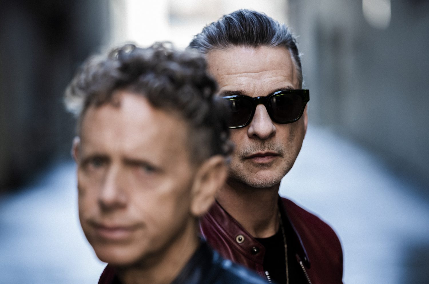 Portrait of two adult males standing close together in a dimly lit urban setting, wearing sunglasses and leather jackets.