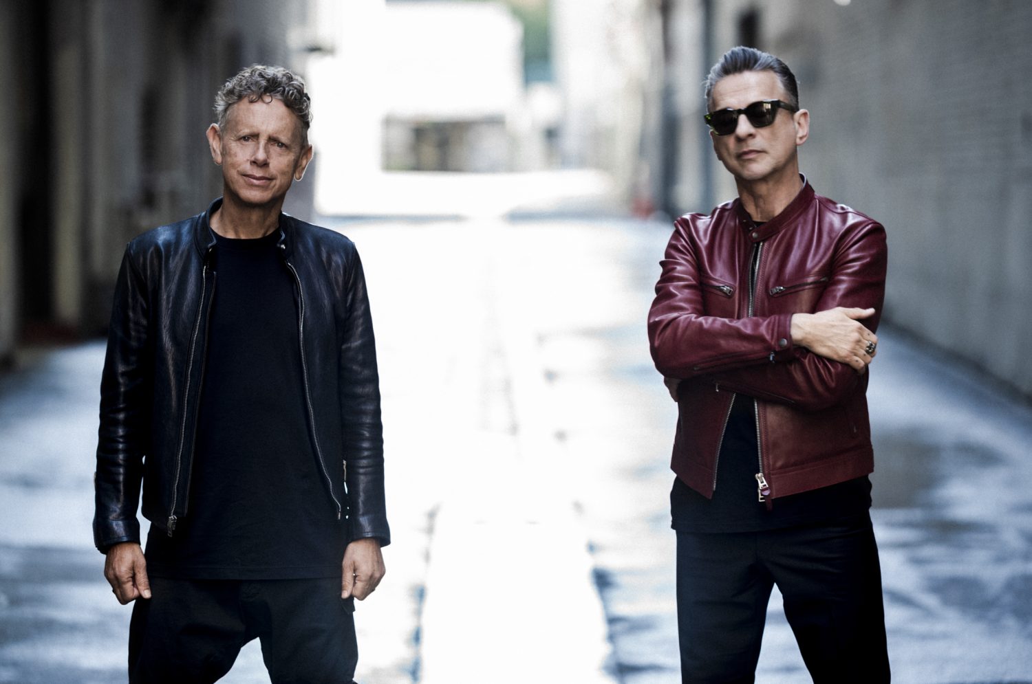 Two adult men stand in a concrete alleyway wearing leather jackets, black shirts, and pants. One wears sunglasses with arms crossed.