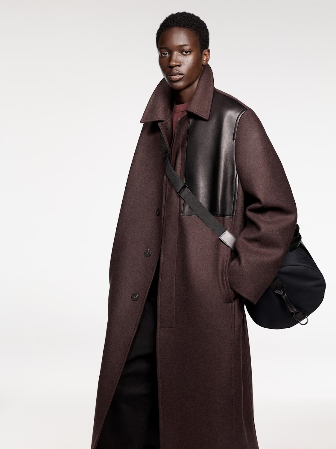Dark-skinned adult model poses in a long, dark red coat with black leather accents and a black bag in a neutral studio setting.
