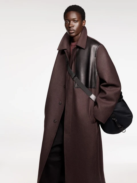 Dark-skinned adult model poses in a long, dark red coat with black leather accents and a black bag in a neutral studio setting.