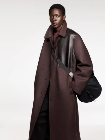 Dark-skinned adult model poses in a long, dark red coat with black leather accents and a black bag in a neutral studio setting.