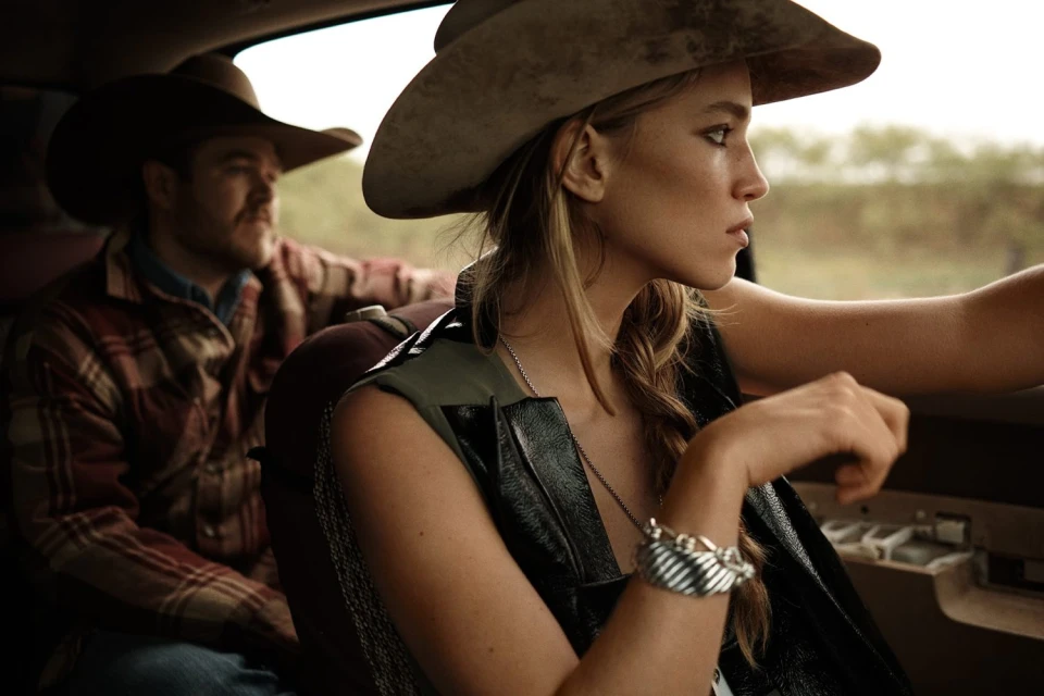 Stylish young woman and man in cowboy hats riding in a vehicle, wearing leather vests and plaid, set against an outdoor rural backdrop.