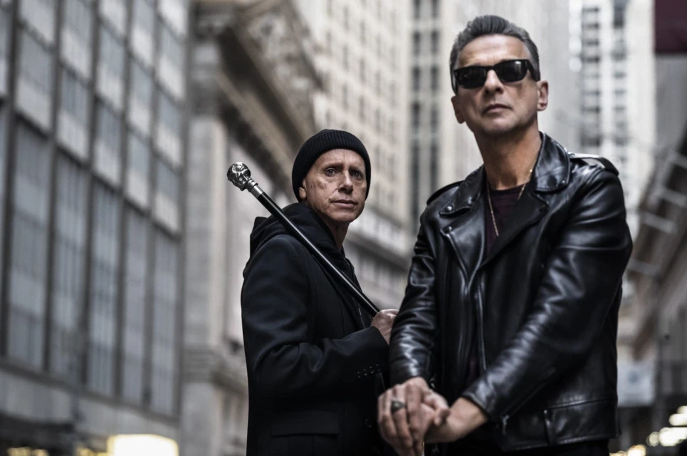 Two men in black outfits stand in an urban environment between tall buildings, one with sunglasses and a leather jacket.
