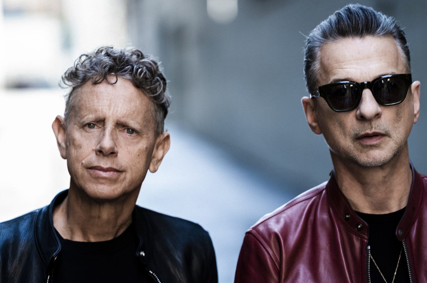 Close-up portrait of two adult males, one with sunglasses and a red leather jacket, the other in a black jacket, against a blurry background.
