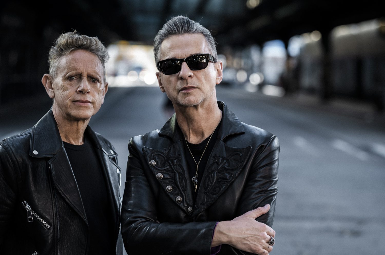 Two adult men stand on a city street in black jackets and sunglasses under an overpass with blurred background in medium shot.