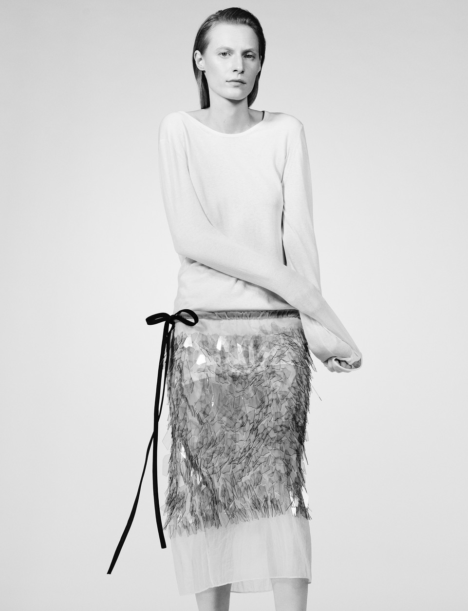 A woman stands in a photo studio wearing a long-sleeved shirt and decorative skirt, presented in black and white with soft lighting.