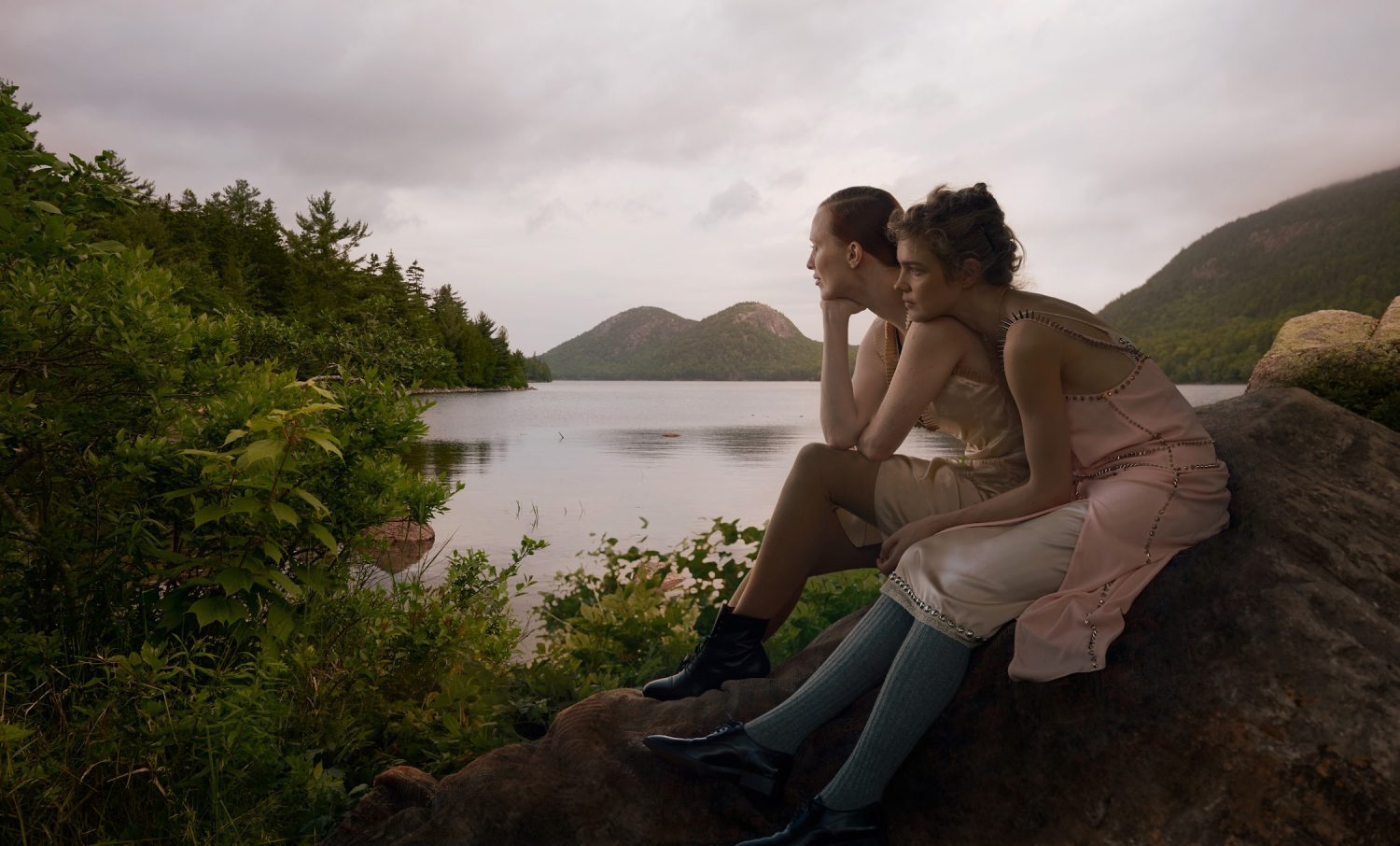 Two women in dresses sit on a rock near a lake surrounded by trees and mountains under a cloudy sky, creating a serene natural scene.