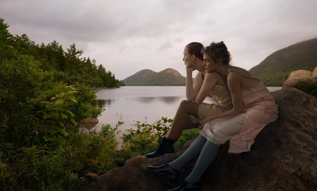 Two women in dresses sit on a rock near a lake surrounded by trees and mountains under a cloudy sky, creating a serene natural scene.