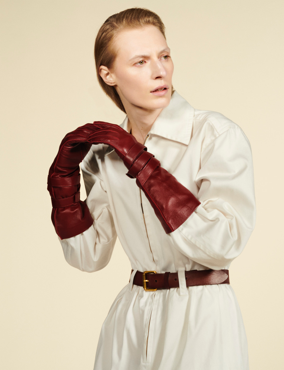 A fashion shot features a fair-skinned adult woman in a studio setting, wearing long burgundy gloves, a white shirt dress, and a brown leather belt.