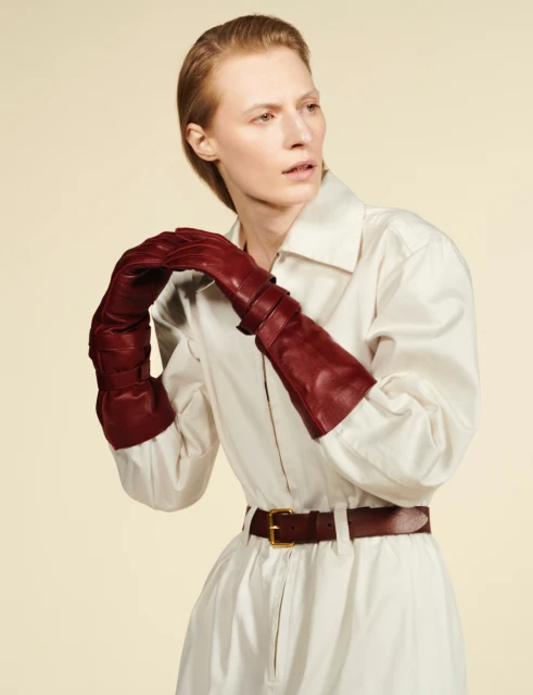 A fashion shot features a fair-skinned adult woman in a studio setting, wearing long burgundy gloves, a white shirt dress, and a brown leather belt.