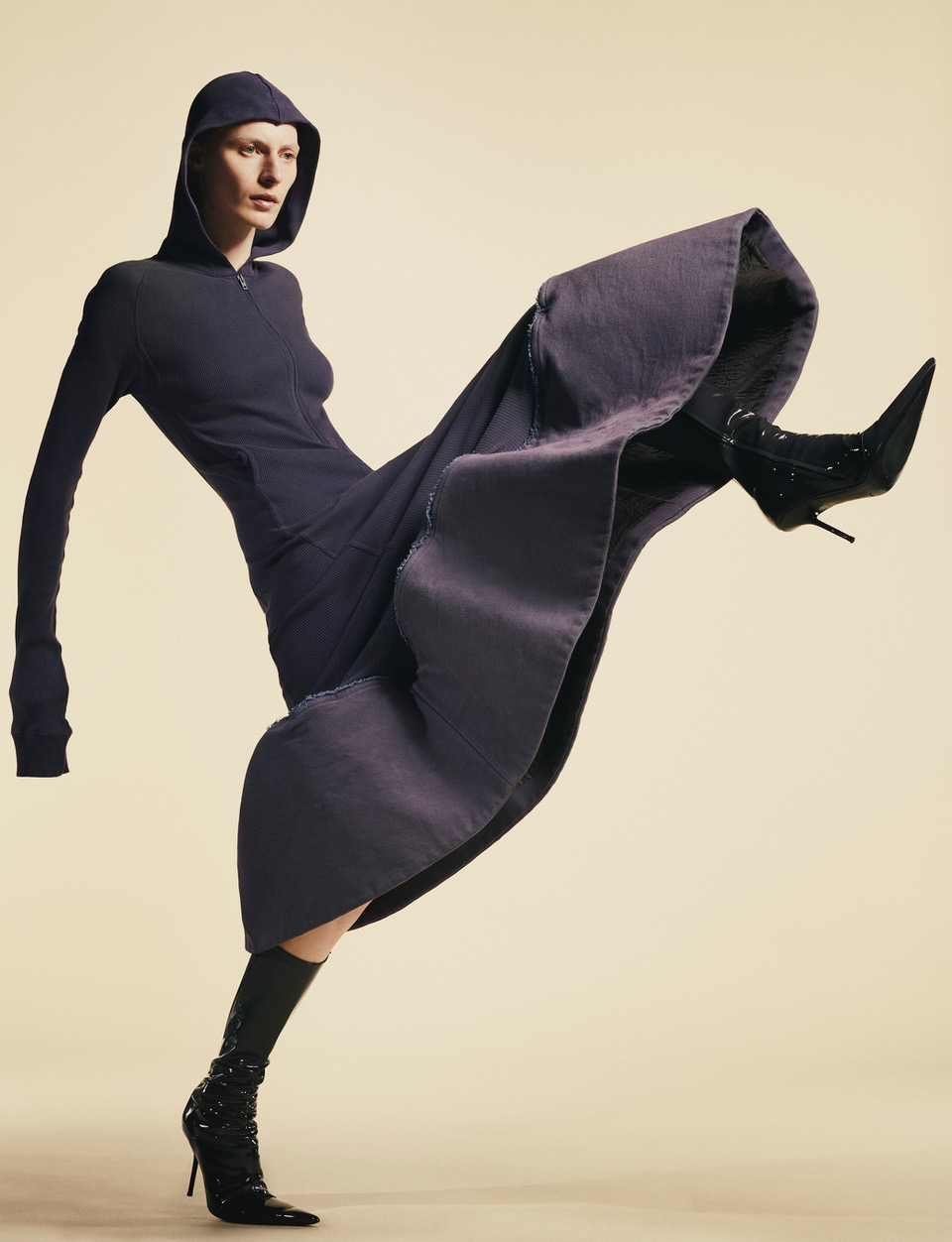 Fashion model in a hooded dress and high-heeled boots strikes a dynamic pose against a neutral backdrop in a studio setting.