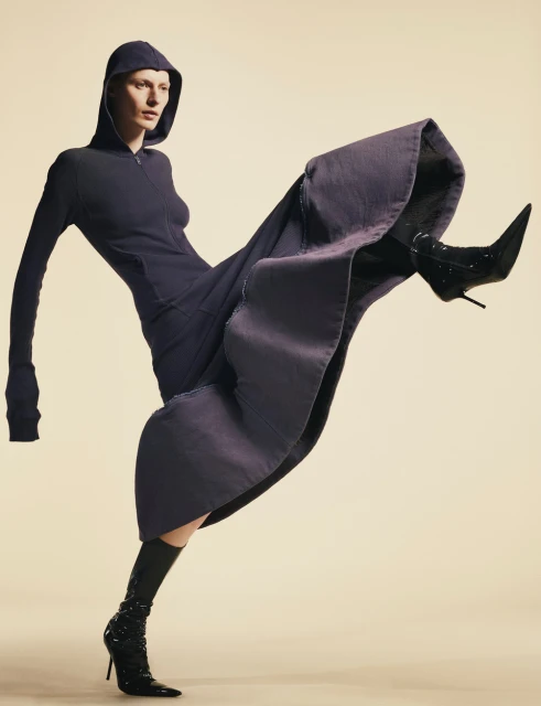 Fashion model in a hooded dress and high-heeled boots strikes a dynamic pose against a neutral backdrop in a studio setting.