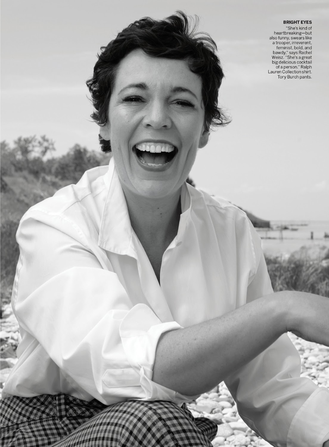 Black and white portrait of a laughing woman wearing a white collared shirt and plaid pants outdoors near water.